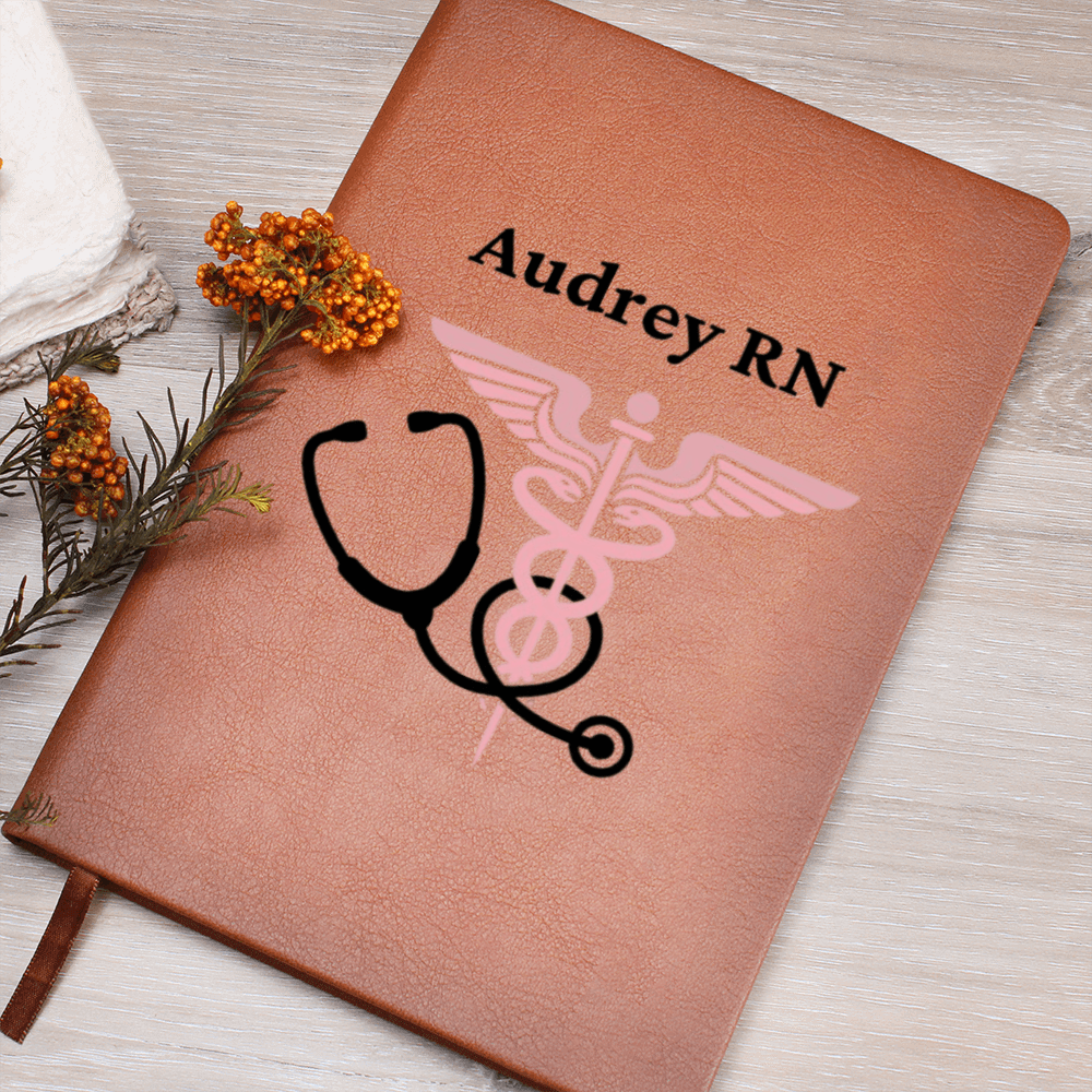 Personalized vegan nurses leather journal with the name 'Audrey RN' and a pink caduceus symbol with a black stethoscope design on the cover, displayed on a wooden surface with floral accents.