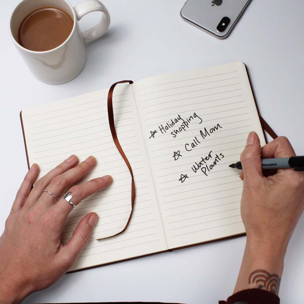 Open lined journal with a brown ribbon bookmark, featuring a handwritten to-do list that includes 'Holiday shopping,' 'Call Mom,' and 'Water plants.' The scene includes a person's hands, a cup of coffee, and a smartphone on a white surface.