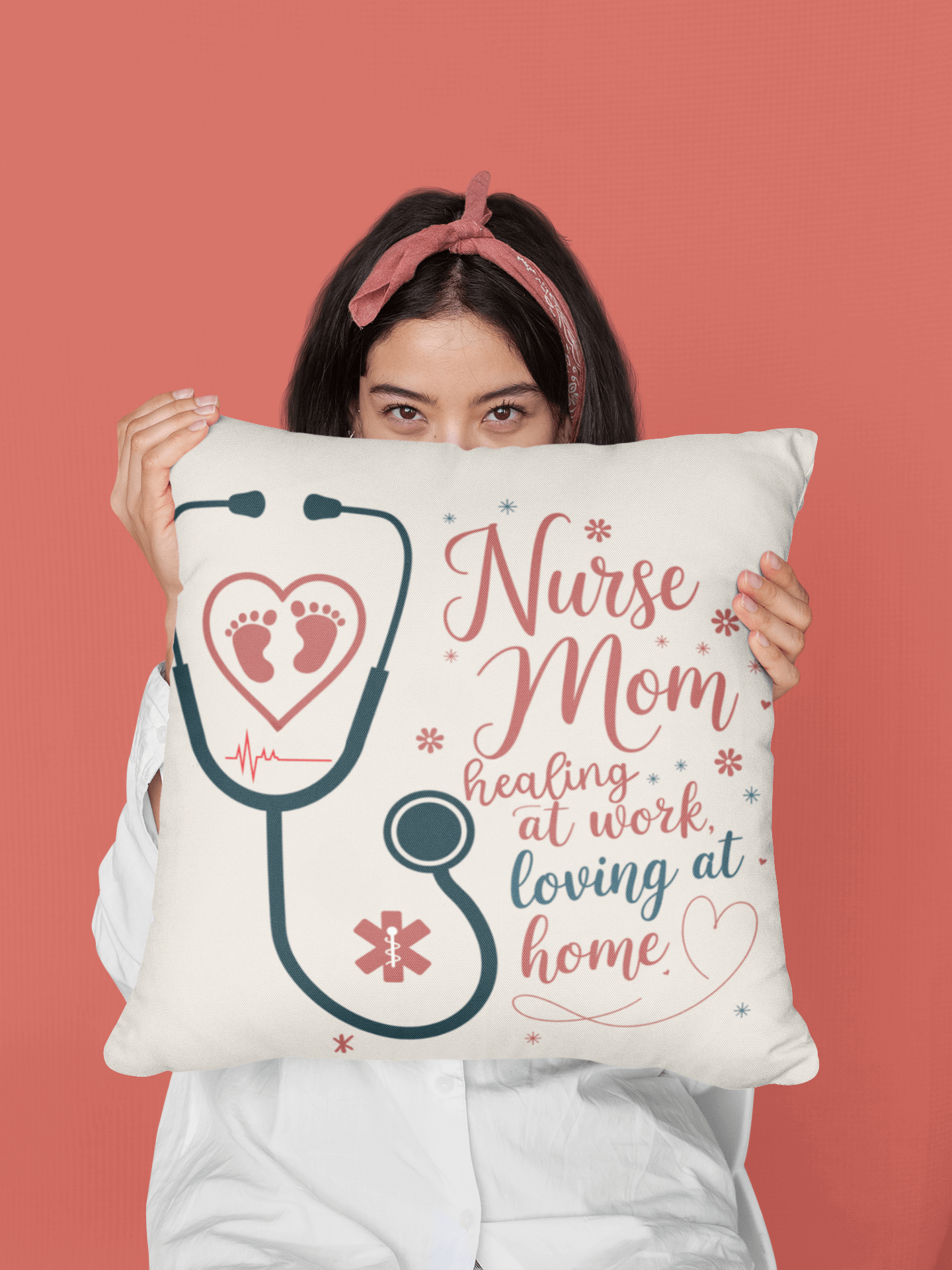 A woman with dark hair and a pink headband holds a nurse-themed pillow in front of her face. The pillow features a stethoscope forming a heart, baby footprints, and the text "Nurse Mom: Healing at work, loving at home" on a warm coral background.