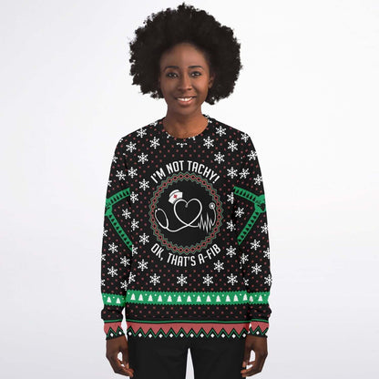 A curly-haired individual is beaming while wearing the "Ugly Christmas Sweater for Nurses – 'I'm Not Tachy! Ok, That's A-Fib' Holiday Sweater," which features a black base adorned with white snowflakes and green and red geometric patterns. The sweater showcases a heart design with the text, "I'M NOT TACHY OK, THAT'S A-FIB." Exuding holiday cheer, they stand against a plain background.