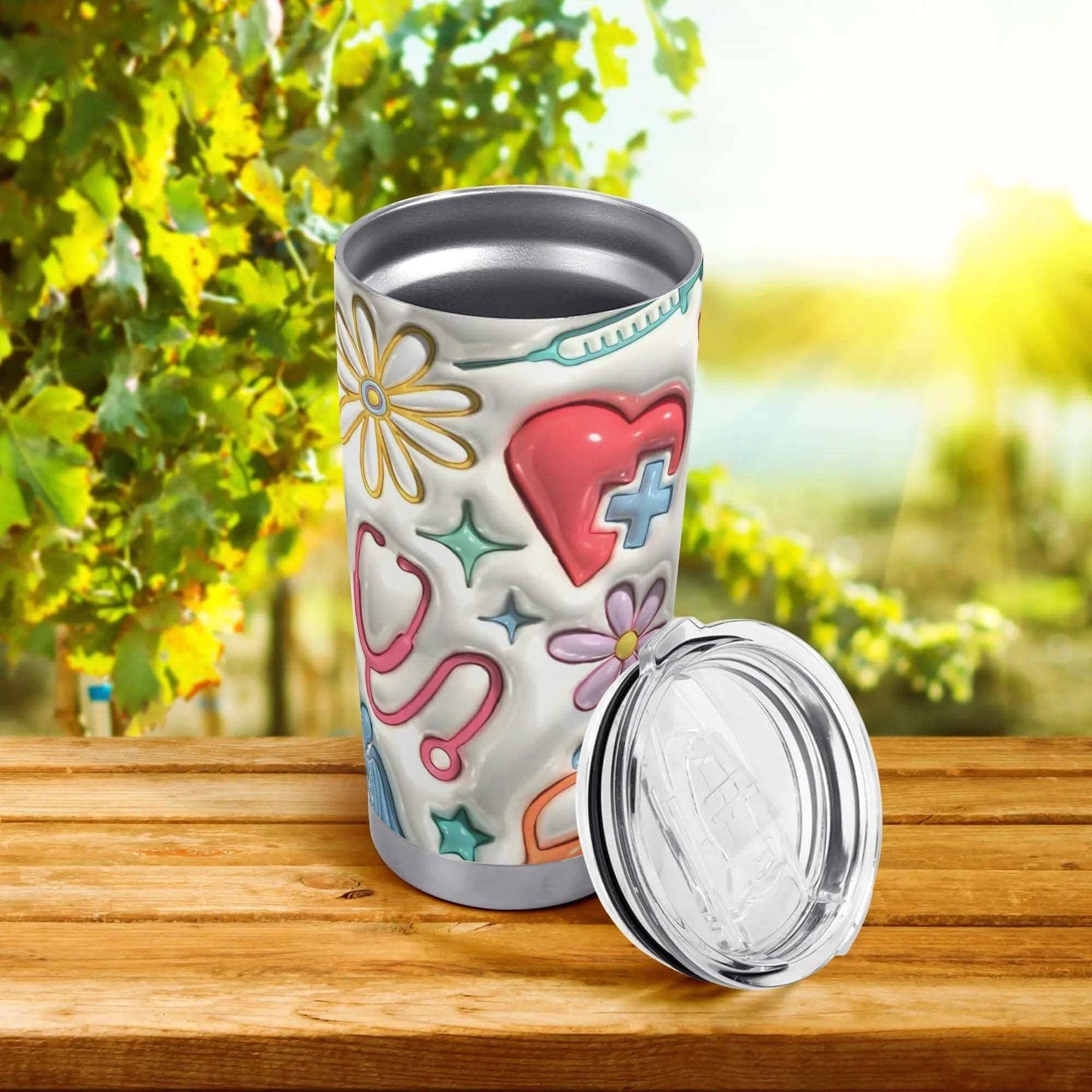 The 20oz 3D Inflated Tumbler - Perfect for Hot & Cold Drinks On-the-Go, adorned with a colorful medical theme design featuring stethoscopes, hearts, and flowers, sits on a wooden surface with its splash-proof lid placed to the side. In the background, a vineyard basks in bright sunlight. The double-walled stainless steel construction ensures your drink remains at the perfect temperature.