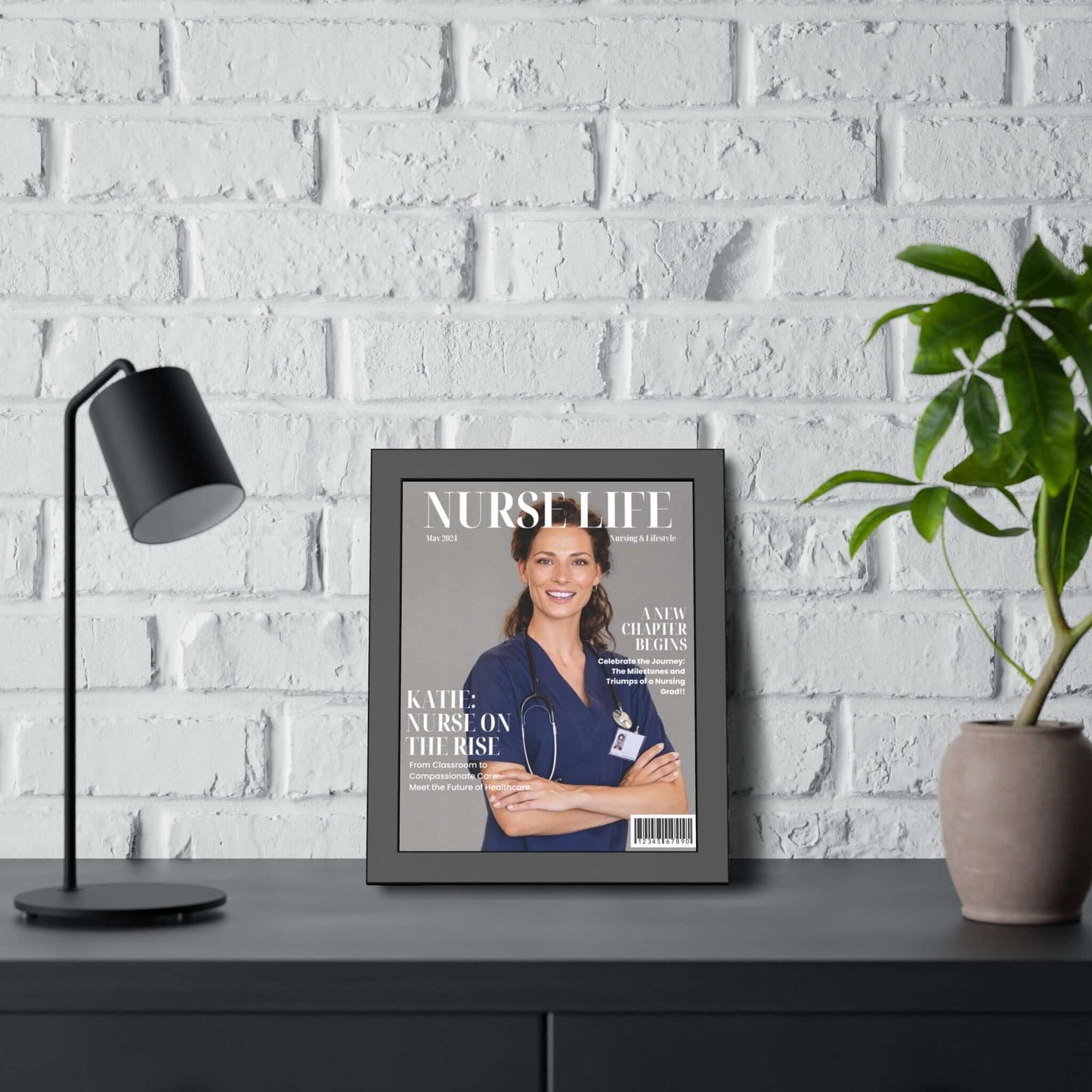 A framed "Personalized Nurse Magazine Cover – Registered Nurse Graduation Gifts" is displayed on a gray desk against a white brick wall. The cover features a smiling nurse in blue scrubs. A black desk lamp is on the left, and a potted plant in a beige pot sits on the right side of the desk—truly a unique and thoughtful gift.