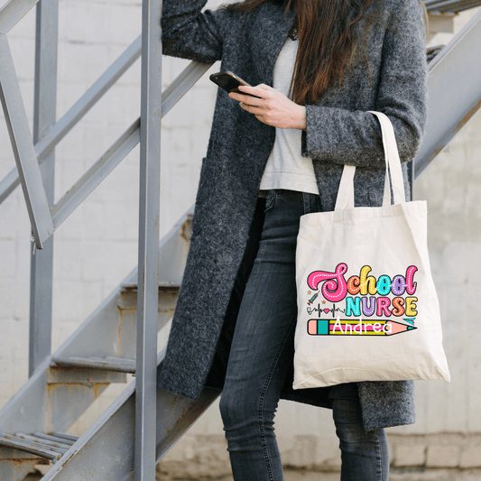 A person wearing a grey coat and jeans stands outdoors on a metal staircase, looking at their phone. They carry a Personalized 100% Cotton Tote Bag for School Nurses with colorful text that reads "School Nurse" and the name "Andrea.