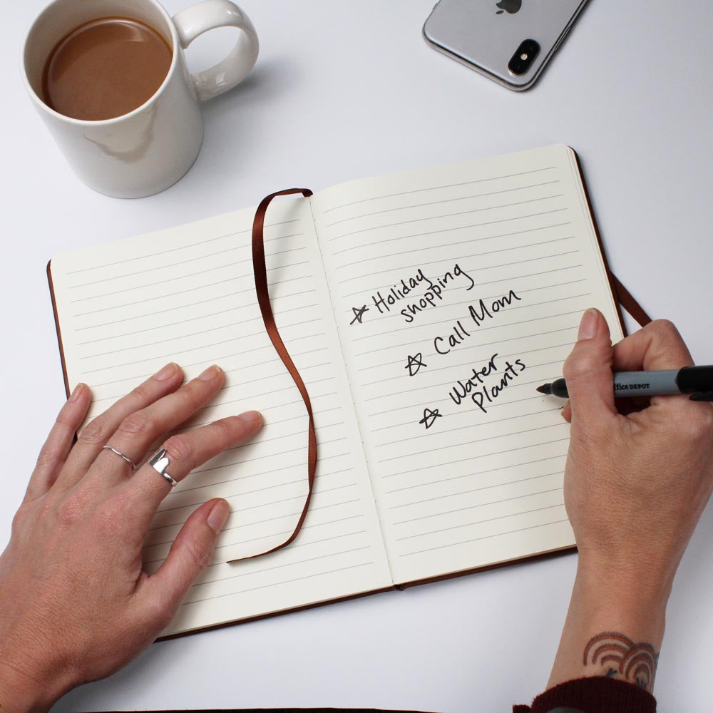 A person's hands are seen writing in an open Custom Medical Journal: Personalized Notebook for Doctors & Nurses - Leather with a black pen. The open page displays a to-do list: "Holiday shopping," "Call Mom," and "Water plants." A cup of coffee, a smartphone, and a pen lie on the white table beside this unique gift.