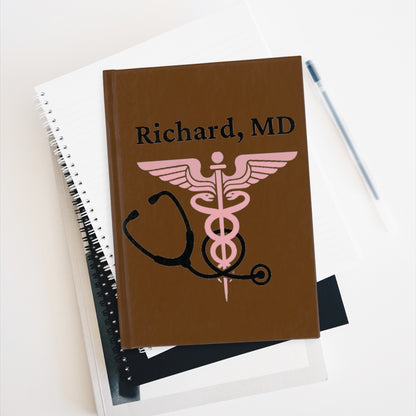 A Custom Medical Journal: Personalized Notebook for Doctors & Nurses with the name "Richard, MD" and a pink caduceus symbol on the cover sits atop a stack of white and black notebooks. A pen lies beside the custom medical journal on a flat white surface.