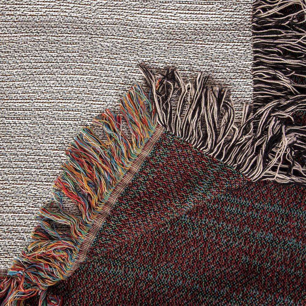 Close-up image of the corner of a woven textile with fringe. The fabric features multicolored threads in shades of red, green, and blue. Another piece of fabric with neutral tones and black and white fringe is partially visible in the background, perfect for a Personalized Nurse Heirloom Woven Blanket - Custom Keepsake for Healthcare Heroes.