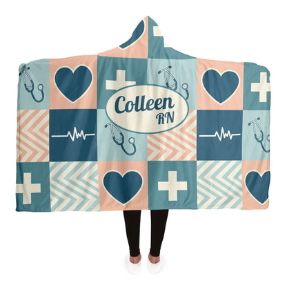 A person wearing a Personalized Nurses Hooded Blanket featuring a patchwork design. The blanket displays teal, navy, and peach squares with symbols such as hearts, crosses, stethoscopes, and EKG lines. Perfect as a nurse gift offering comfort and relaxation, it has "Colleen RN" printed in a central oval.